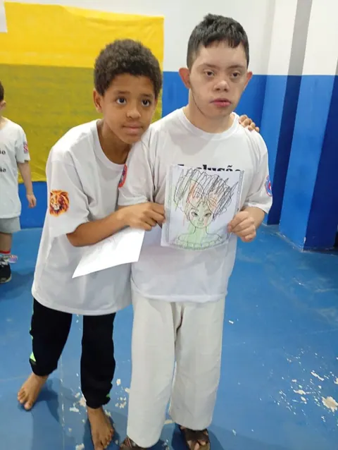 Dois alunos de kimono abraçados, um deles mostrando um desenho